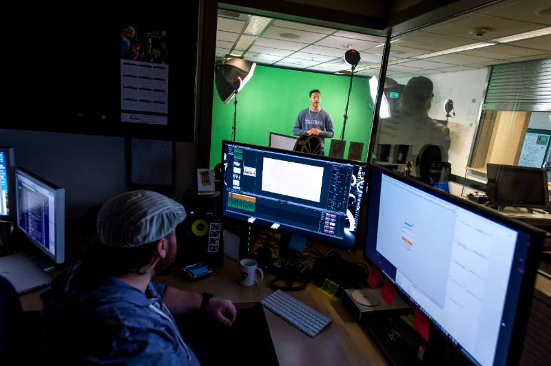 A student stands in front a green screen and video set up, while another edits footage on multiple screens