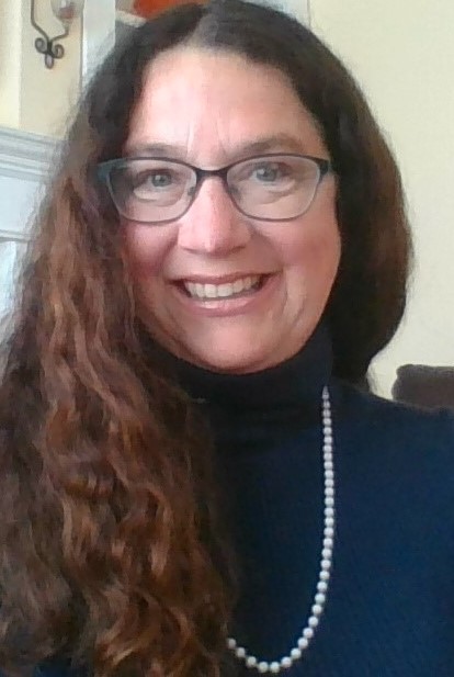 A woman with long brown hair wearing glasses, a sweater and a necklace. 