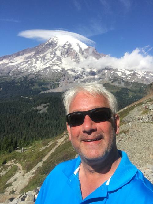 Picture of a man with white hair and sunglasses in front of a mountain. 