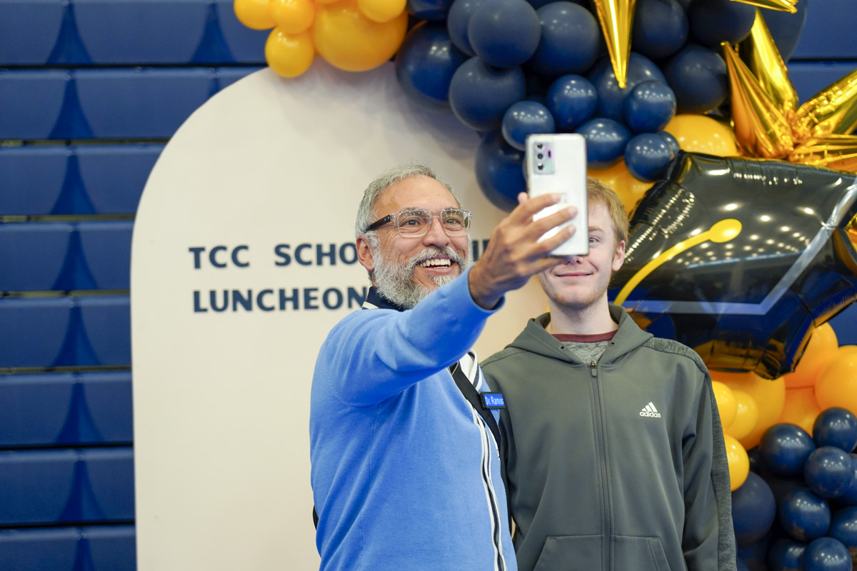 A man with a beard and glasses smiling as he talkes a selfie with a man in a green hoodie. 