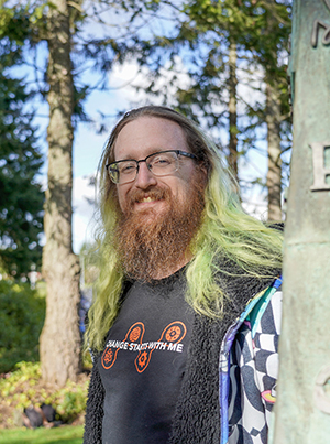 A man with green hair and a beard by the TCC peace pole smiling. 